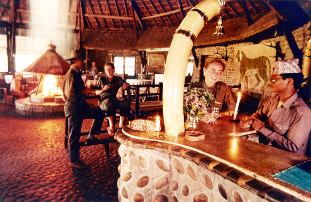 People gather around a stone-faced bar beneath exposed rafters, with a fireplace in the sitting area behind.