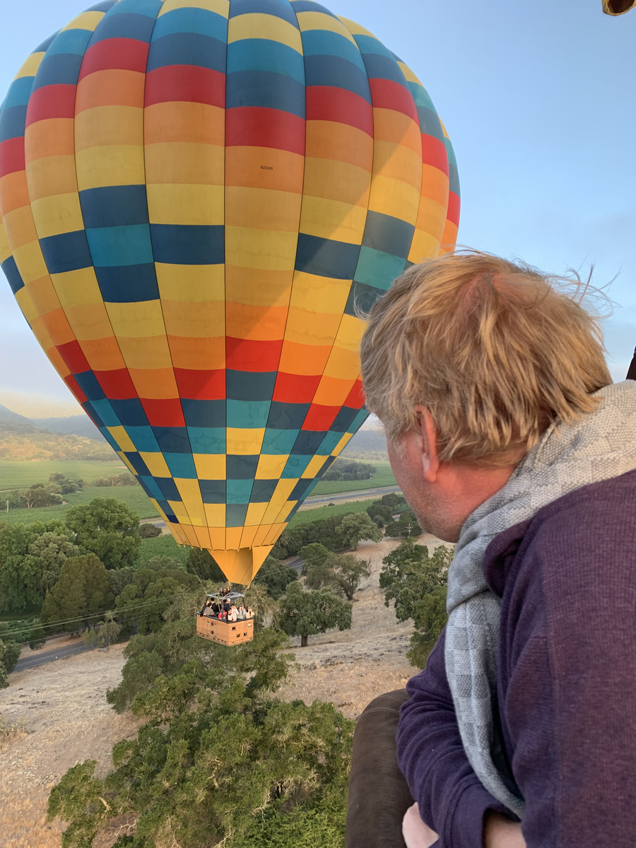 Konstantin looking from a balloon basket towards another colourful balloon over the vineyards.