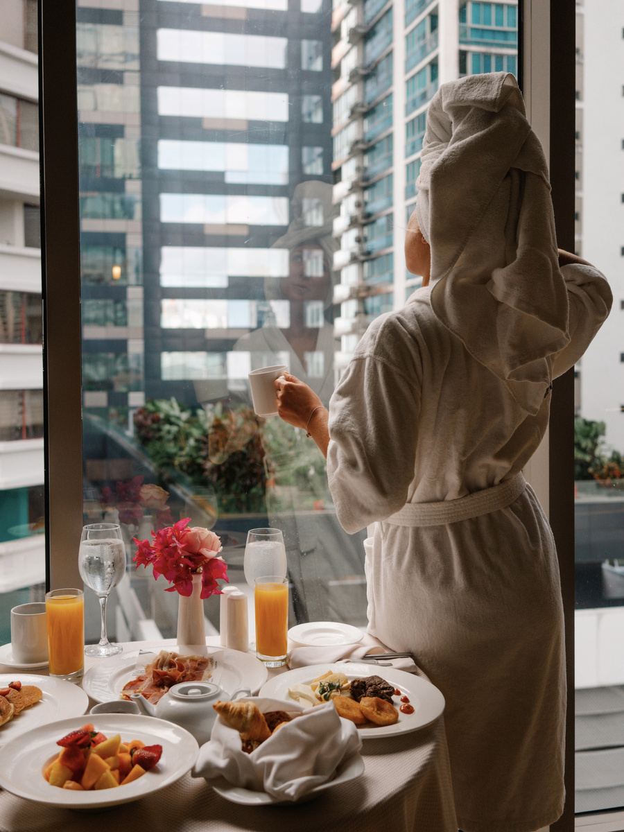 Olga in a bathrobe beside the room-service breakfast table and the window.