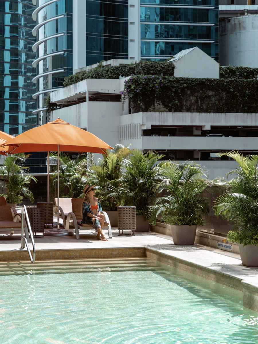 Olga seated under an orange parasol beside the hotel pool, with city towers behind her.