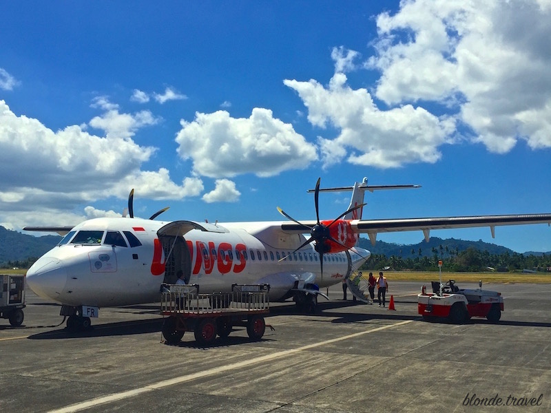 A Wings Air ATR turboprop on the apron of a small Indonesian airport under a bright sky with clouds.